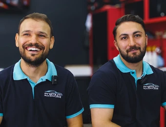 Zwei Männer in dunklen Poloshirts mit hellblauem Kragen, lachen; vor roten Regalen in einer Werkstatt.