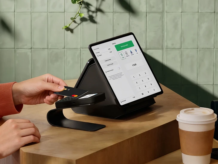 Person hält Karte an Kartenterminal bei POS-Station; Tablet zeigt Kassenschnittstelle; Kaffeebecher auf Holztisch.