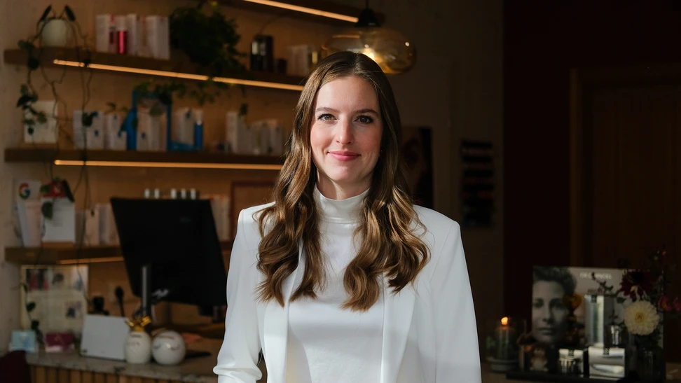 Eine Frau mit braunen, welligen Haaren trägt einen weißen Blazer und weißen Rollkragen und steht vor Regalen in einem Laden.