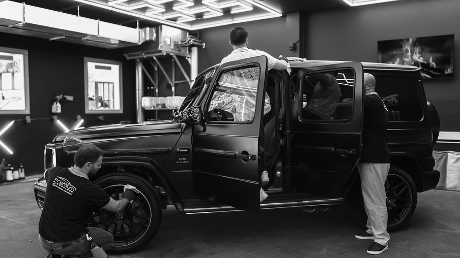 Schwarzer SUV in einer Werkstatt; drei Personen arbeiten am Fahrzeug, Tür geöffnet.